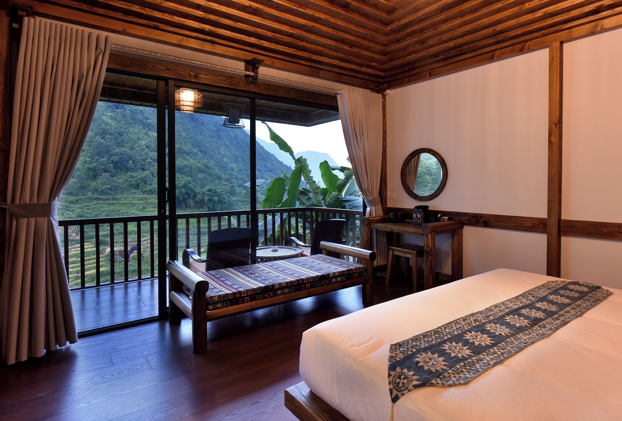 A bedroom with a bed, a desk, a chair, and a view of a lush landscape through large windows.
