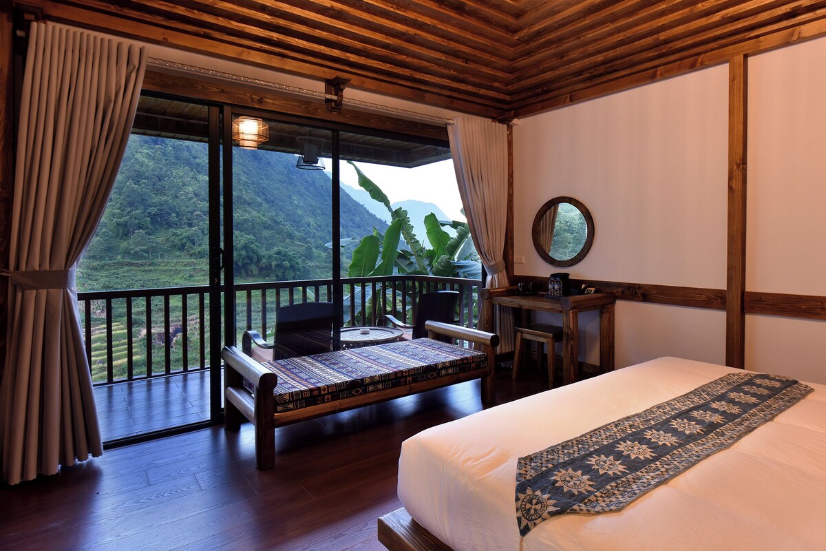 Quarto com cama, escrivaninha, cadeira e vista para uma paisagem exuberante através de grandes janelas.