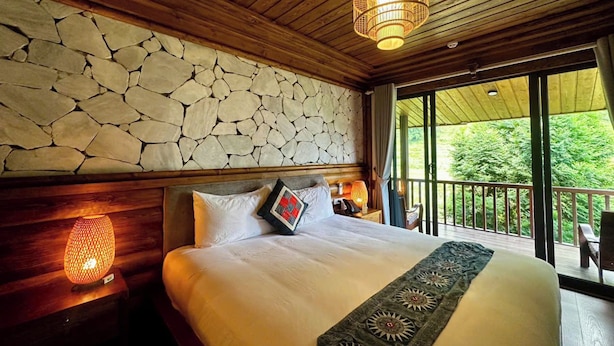 A bedroom with a stone wall, a wooden ceiling, a bed with white bedding, a nightstand with a lamp, and a view of greenery outside. - MAISON DE LAO CHAI (Sa Pa)