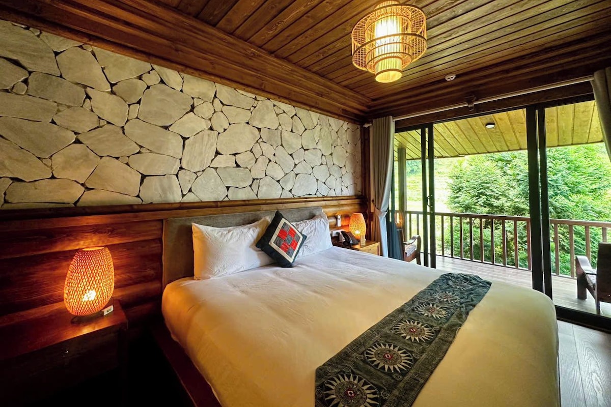 Un dormitorio con pared de piedra, techo de madera, cama con ropa blanca, mesita de noche con lámpara y vistas al exterior lleno de vegetación.