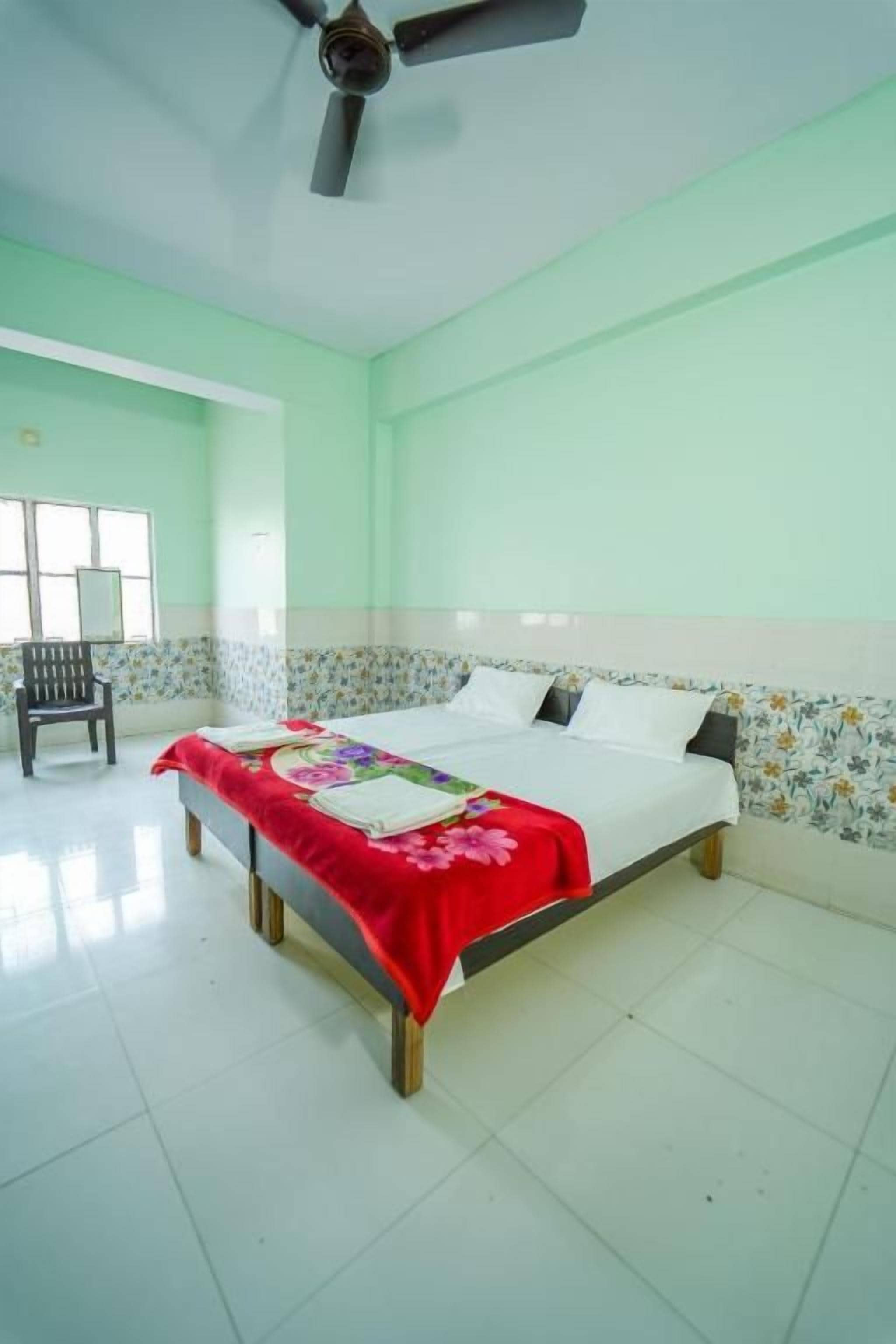 A single bed with a red bedspread, a ceiling fan, and a chair in a room with light green walls and a tiled floor.