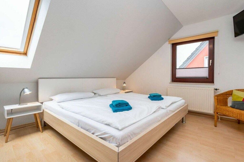 Une chambre avec un plafond en pente, un lit avec une literie blanche, deux serviettes bleues, une lampe de chevet, un fauteuil en osier et une fenêtre avec des stores.