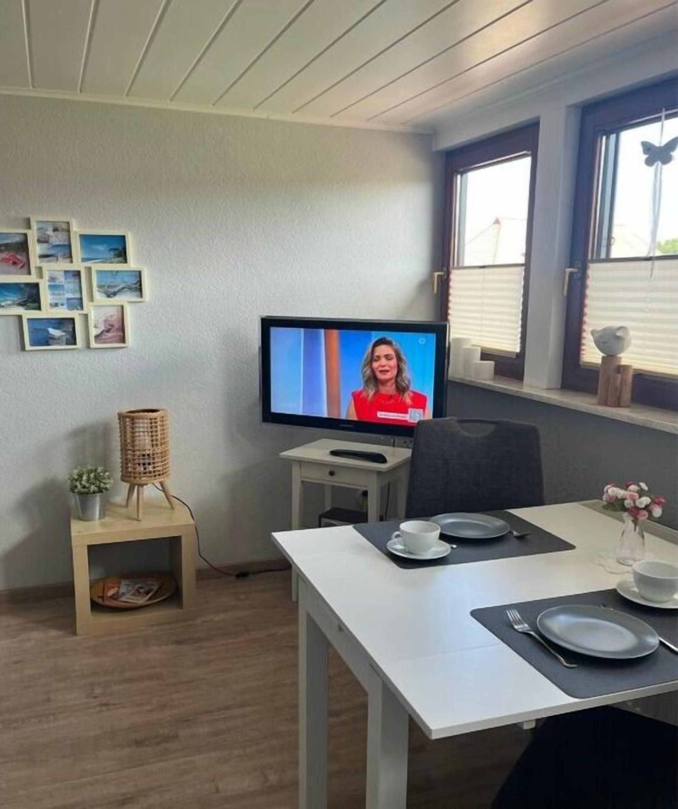 Ein Zimmer mit Fernseher an der Wand, einem gedeckten Esstisch mit Geschirr und Besteck sowie einem kleinen Beistelltisch mit Korb und einem Fenster mit Jalousien.