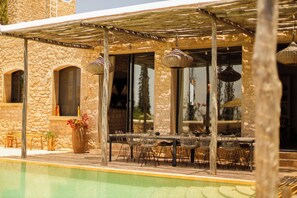 Uma área de refeições ao ar livre com paredes de pedra, piscina e guarda-sóis.