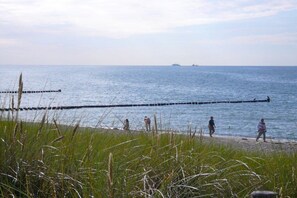 Ein Strand mit einer langen Holzsteganlage, die sich ins Wasser erstreckt, Menschen, die am Ufer entlanggehen, und ein Schiff in der Ferne.