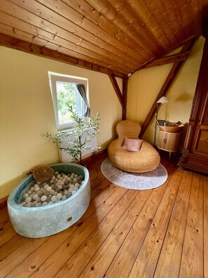 Une pièce chaleureuse avec un plancher en bois, un fauteuil rond en osier agrémenté d’un coussin, une petite table ronde avec une lampe, et un grand récipient rond rempli de balles.