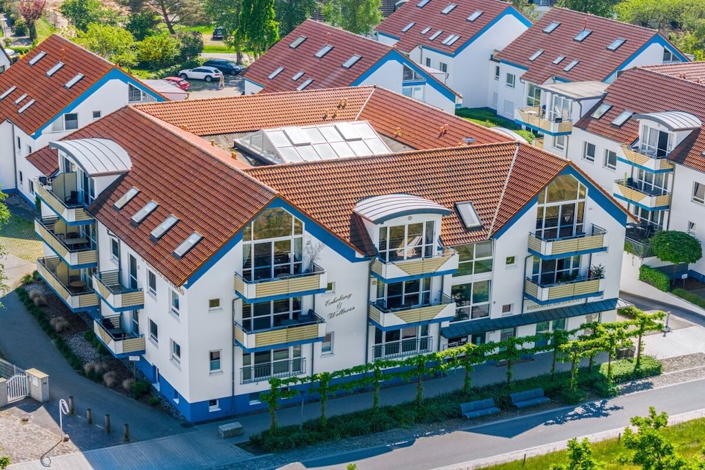 Un immeuble résidentiel à plusieurs étages, doté d’un toit recouvert de tuiles rouges, de balcons et d’un jardin paysager à l’avant.