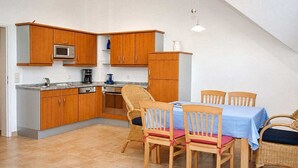 A compact kitchen with wooden cabinets, a microwave, and a dining area with wicker chairs and a blue tablecloth. - Residence Am Strand 1-16 (Seeheilbad Zingst)