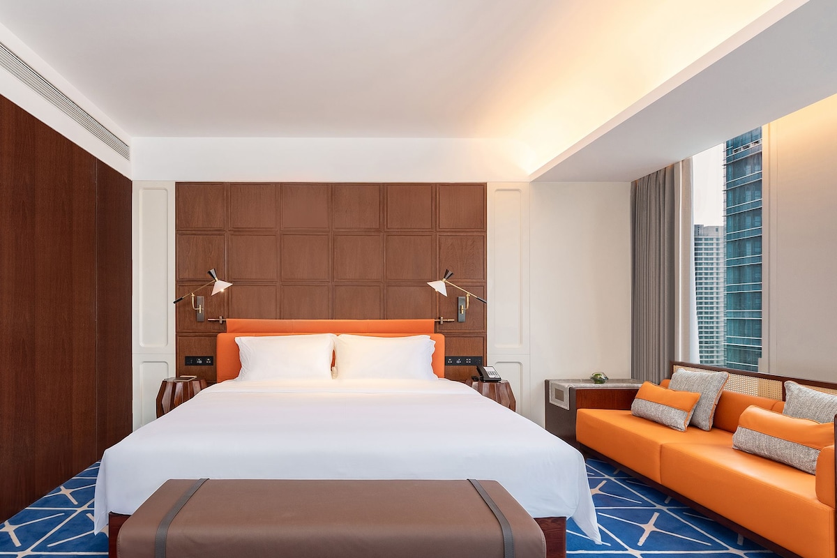 Un cuarto de hotel con una cama grande, un sofá naranja, un banco y vistas de un paisaje urbano.