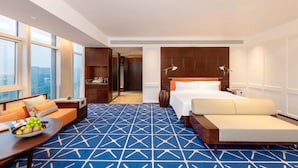Een hotelkamer met een bed, een bank, een eettafel met fruit, een groot raam en uitzicht op de stad.