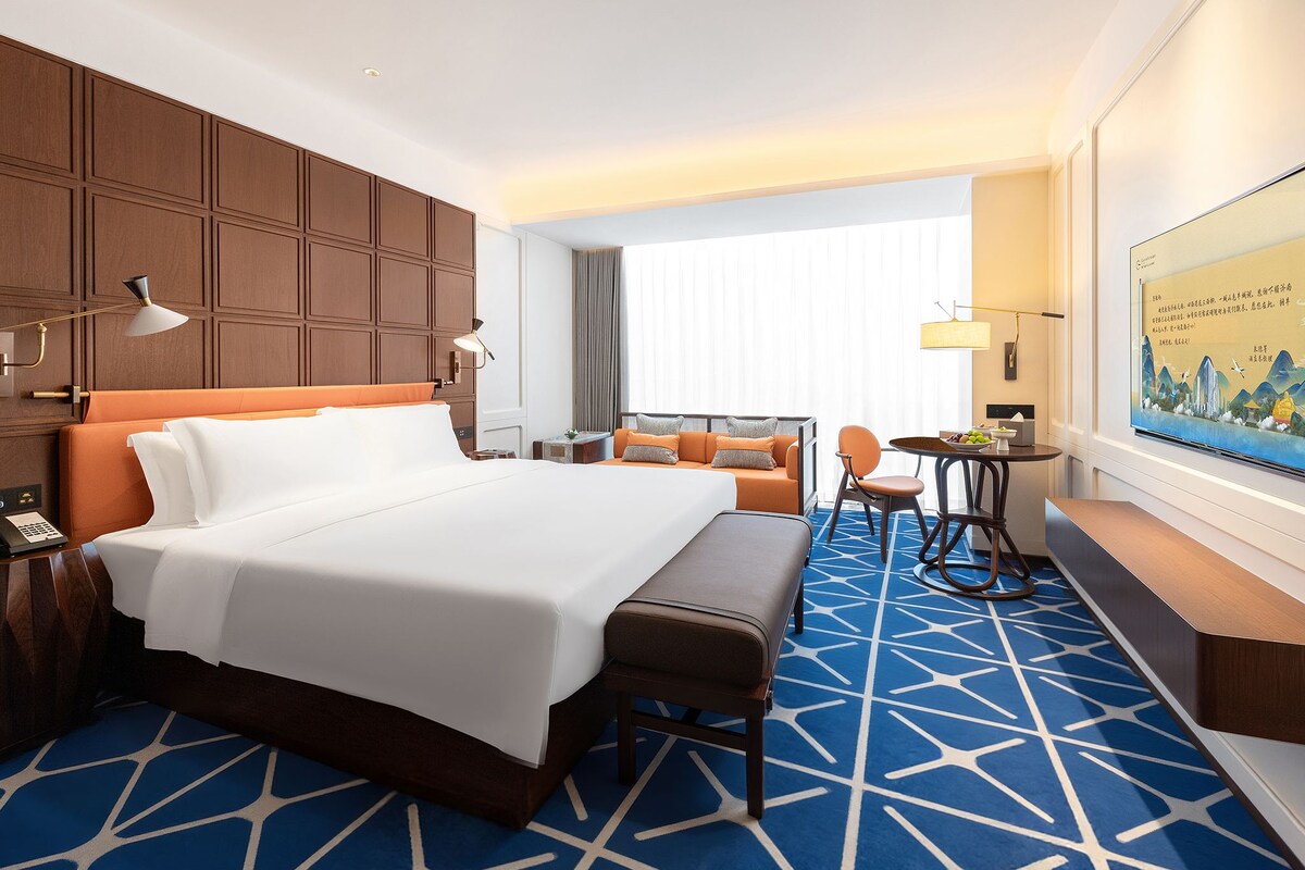 Habitación de hotel con una cama grande, un sofá, una mesita y un banco. Hay un cuadro en la pared y una ventana con cortinas.
