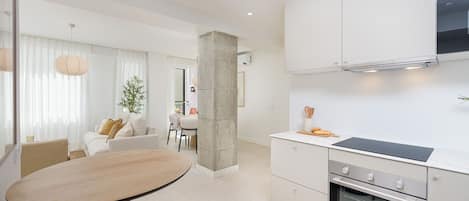 A modern kitchen with a built-in oven, a dining area with a round table, and a living space with a sofa and curtains.
