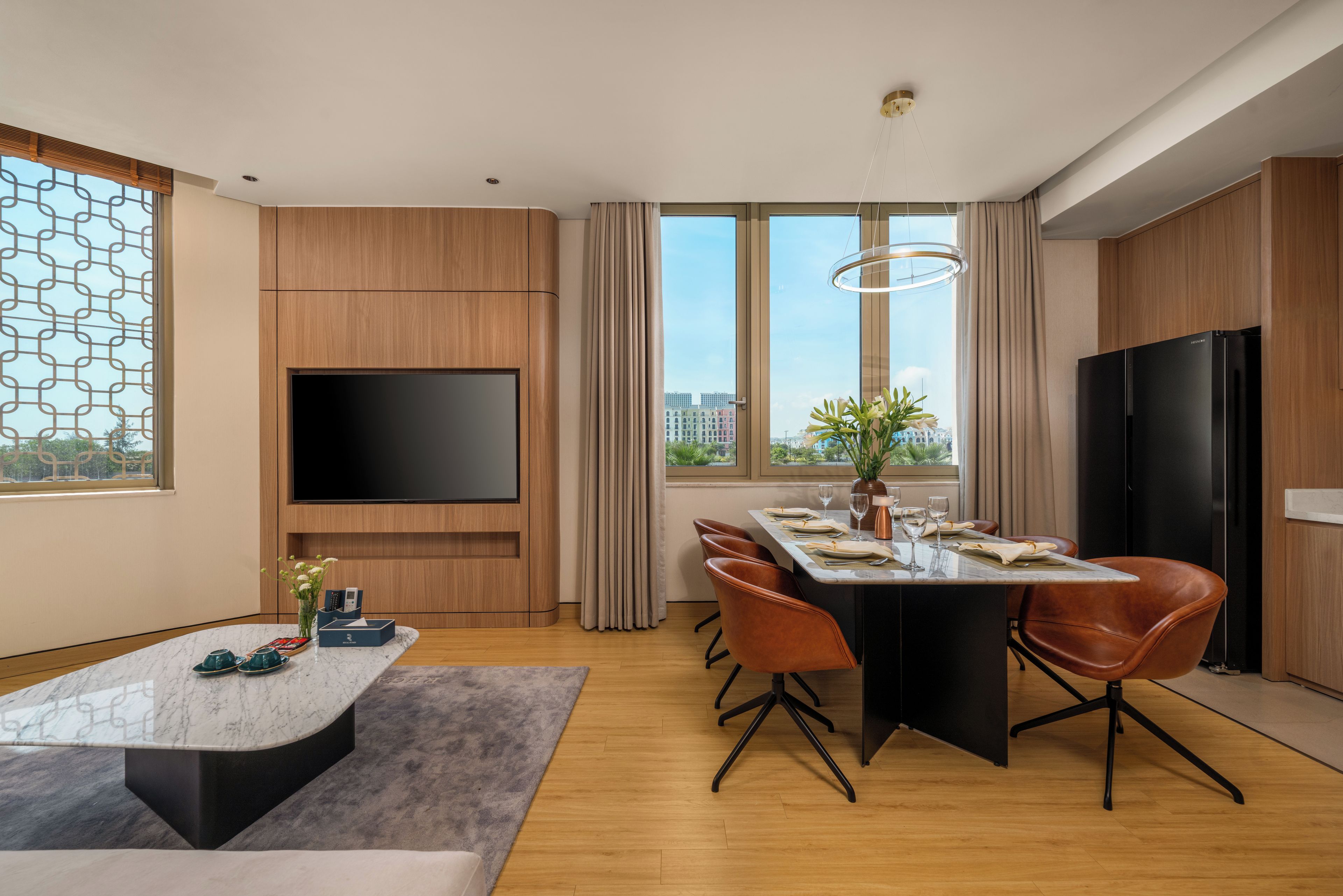 A modern living room with a flat-screen TV, a dining area with a marble table, and a large window offering a city view.