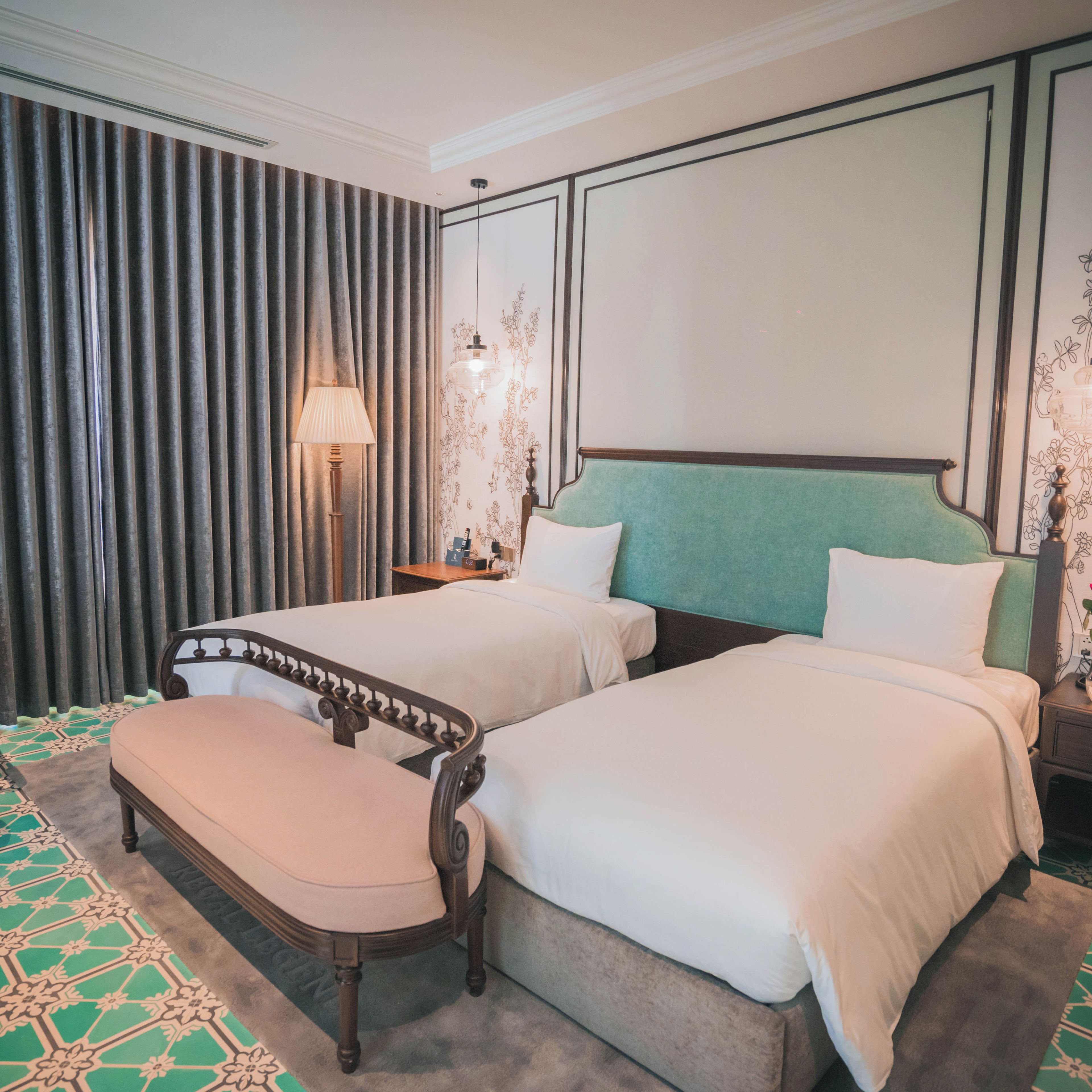 Ein Hotelzimmer mit zwei Betten, einer Bank, einem Nachttisch, einer Lampe und einer Wand mit Blumenmuster.