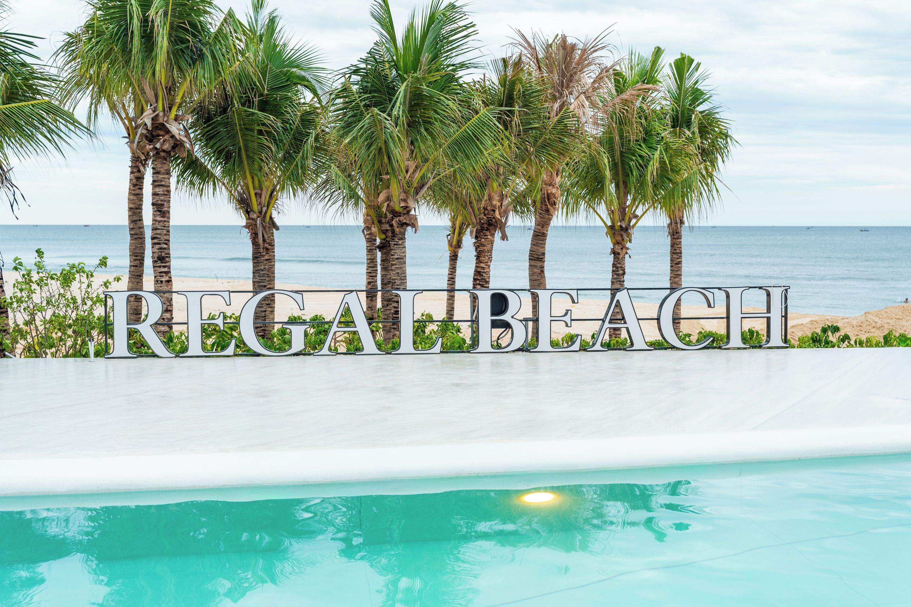 A sign with the text 'REGABEAH' in front of a pool with a view of the beach and ocean.