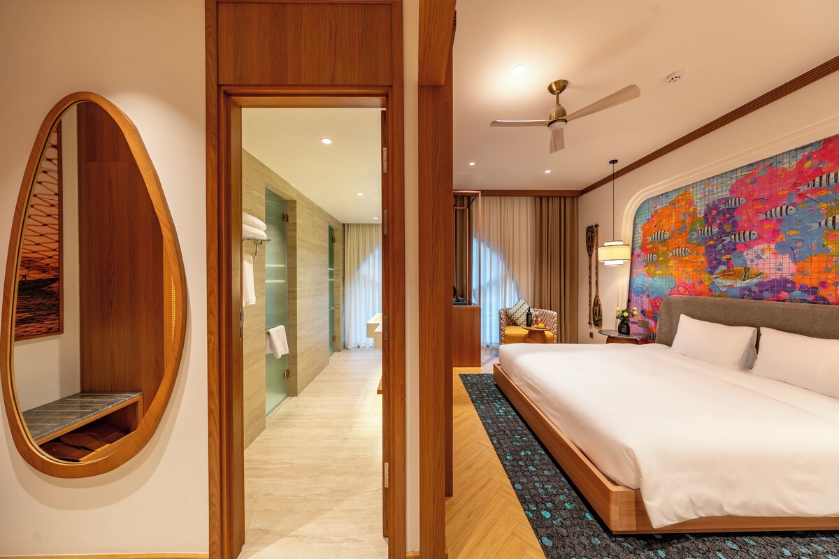 Habitación de hotel con una cama grande, una puerta de madera, un baño con ducha de vidrio y un mural de peces coloridos en la pared.