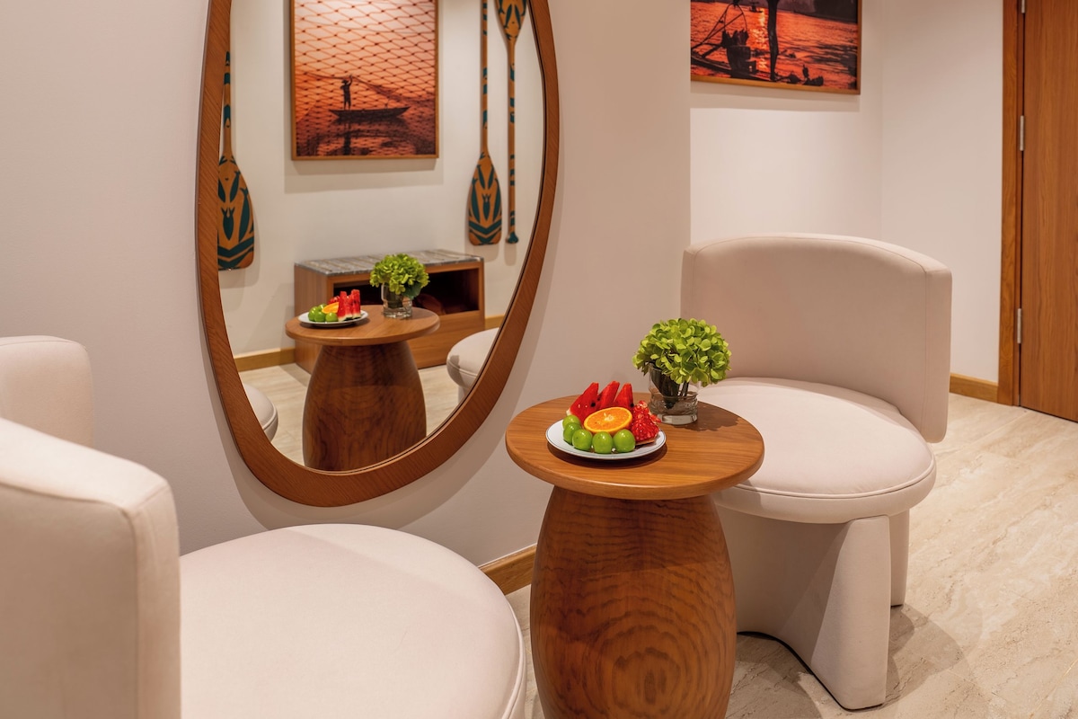 A modern interior with a large oval mirror, two white armchairs, a small wooden table with fruit, and artwork on the walls.