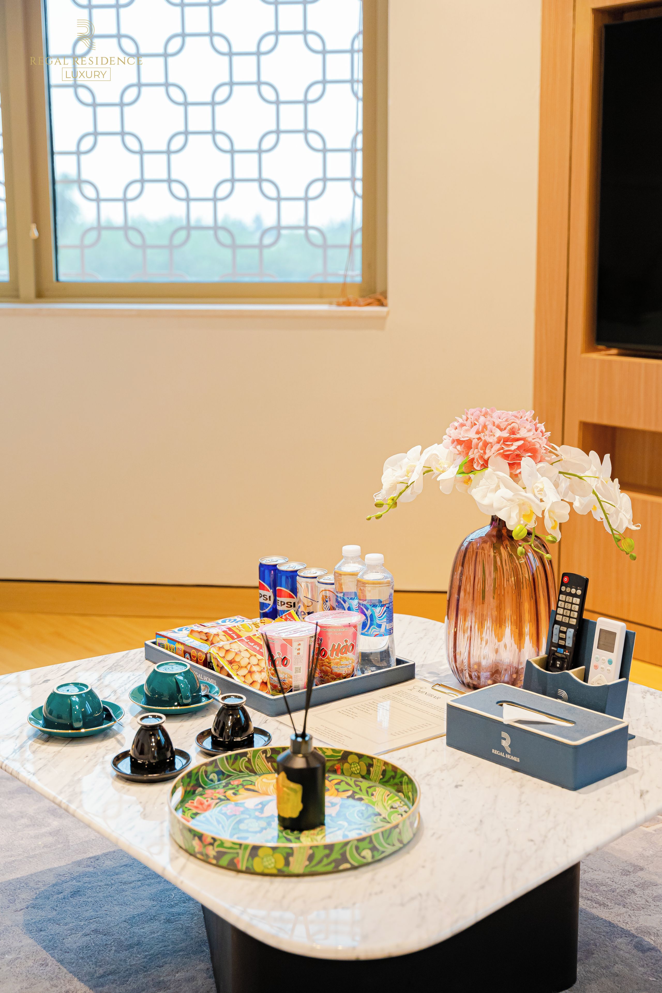 A hotel room with a marble table set with various amenities including a vase of flowers, a tray of snacks, and a remote control.
