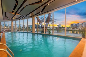 Une piscine intérieure offrant une vue dégagée sur le coucher de soleil et le paysage urbain grâce à de grandes fenêtres.