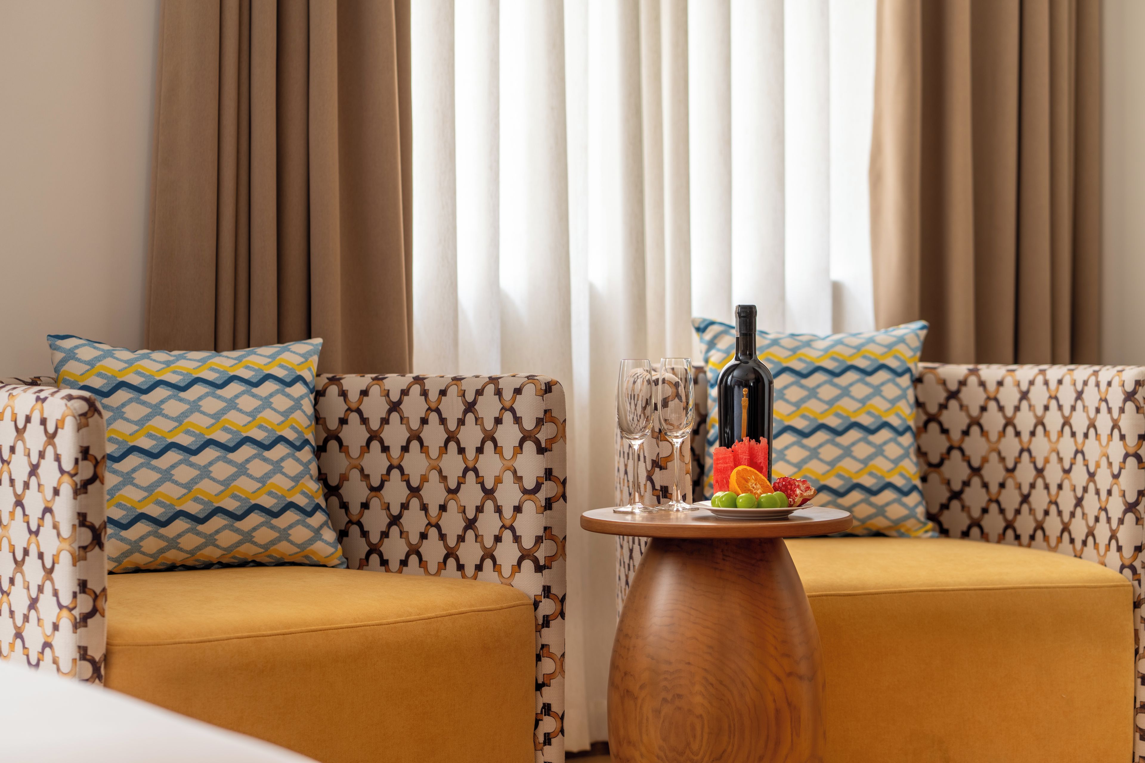 A modern living room with patterned sofas, a wooden side table with a bottle and glasses, and a view through sheer curtains.