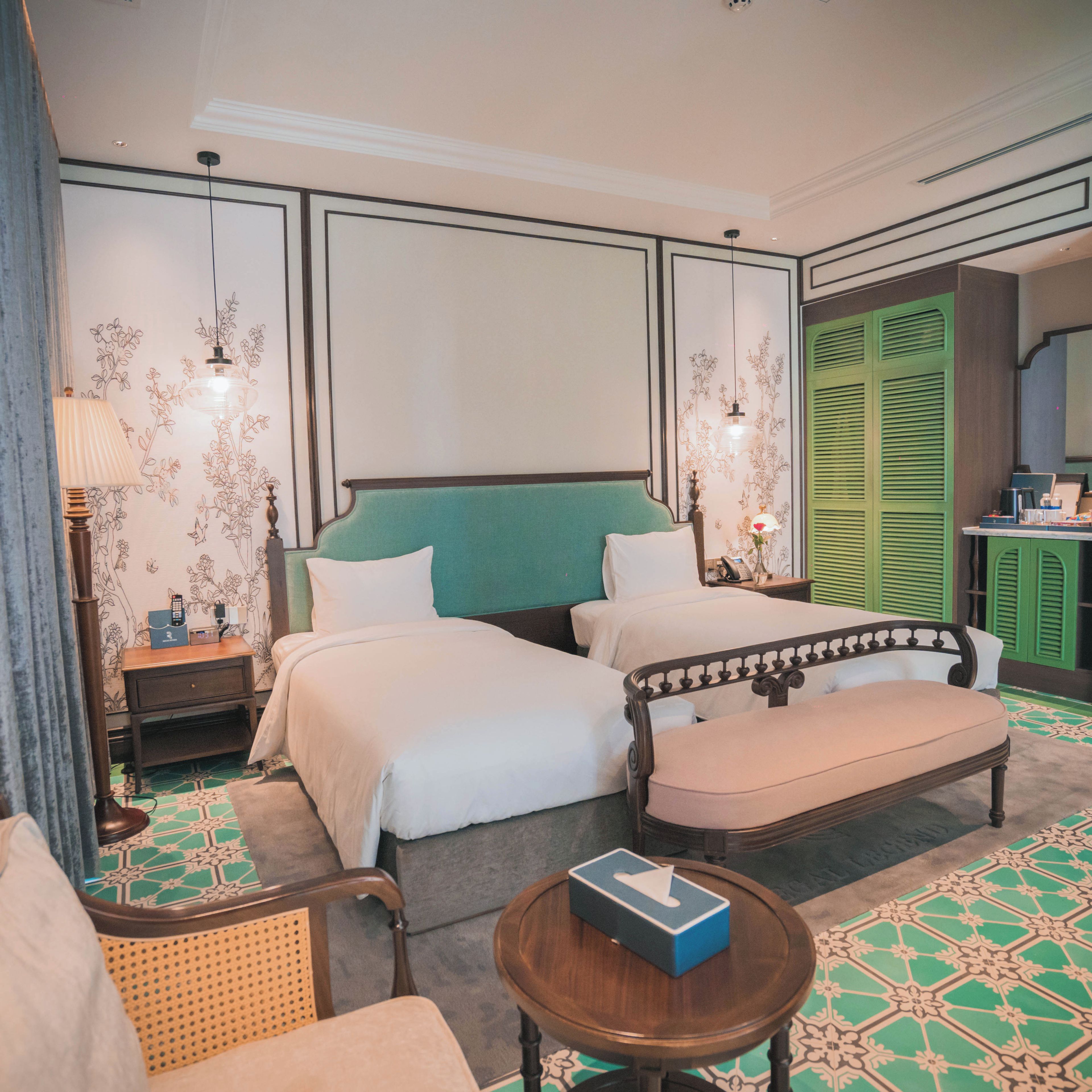 A hotel room with two beds, a green headboard, a wooden nightstand, a bedside table, a chair, and a small round table with a tissue box.