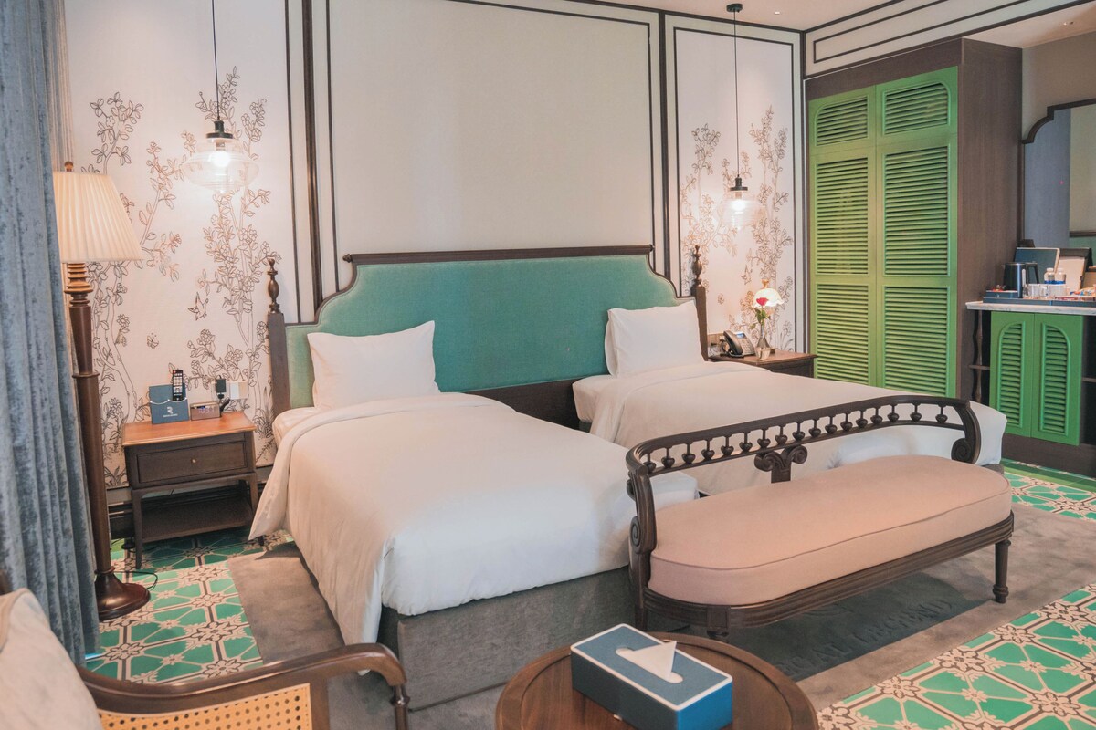 Habitación de hotel con dos camas, un cabecero verde, una mesita de noche de madera, una mesilla auxiliar, una silla y una pequeña mesa redonda con una caja de pañuelos.