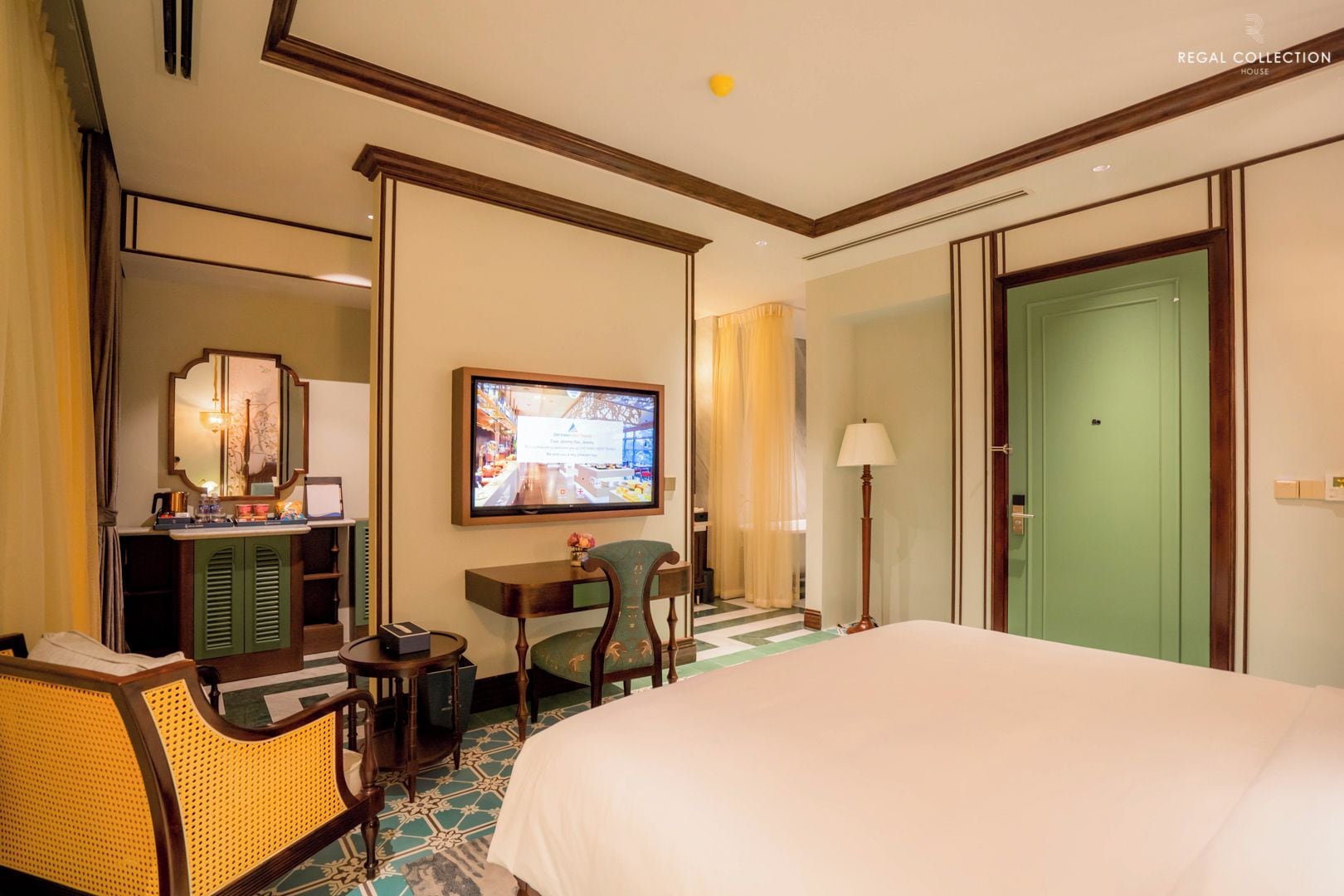 Habitación de hotel con cama, escritorio, televisor, espejo y puerta verde.