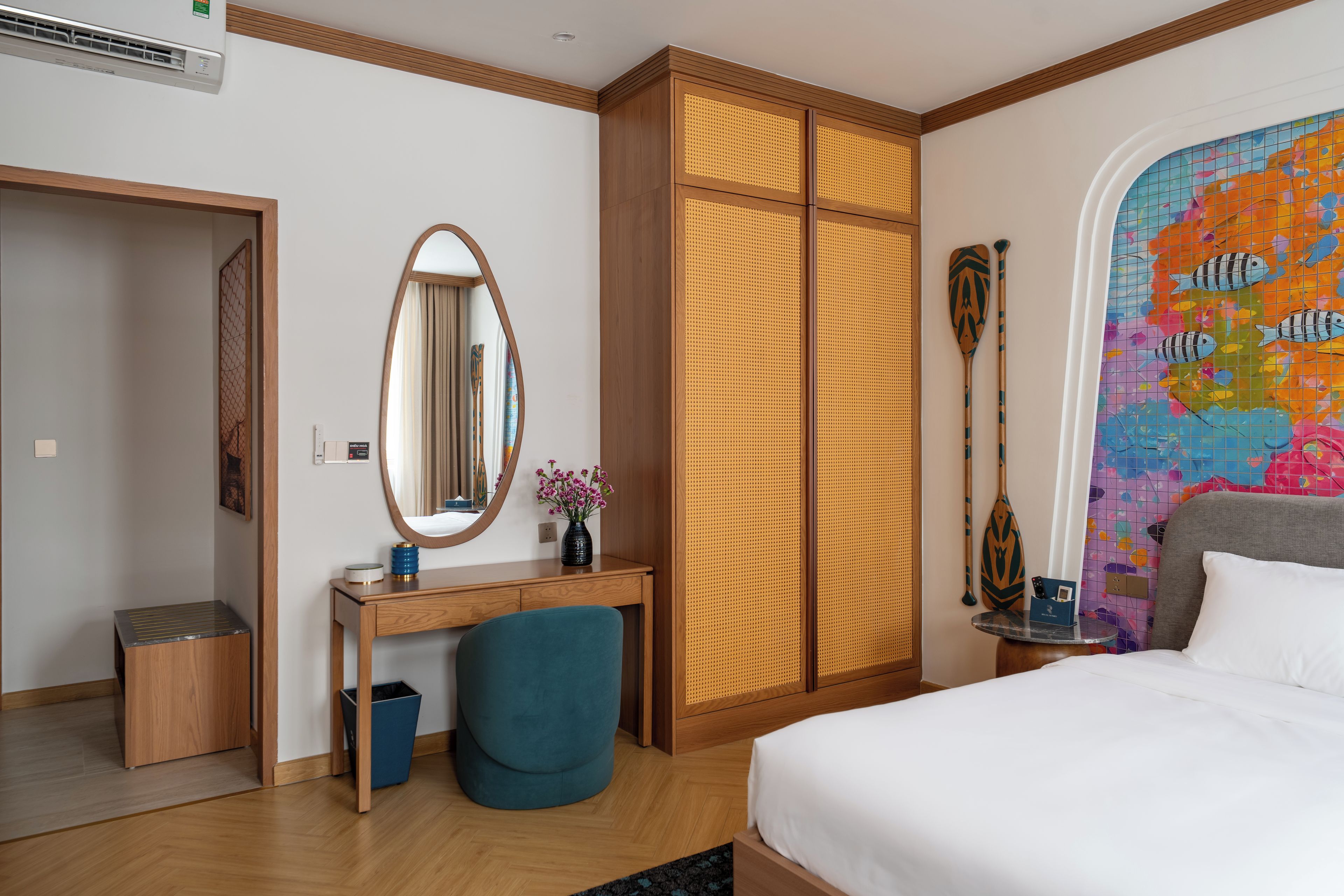 A hotel room with a bed, a desk, a chair, a wardrobe, a mirror, a vase with flowers, a wall with a colorful map, and a window with curtains.