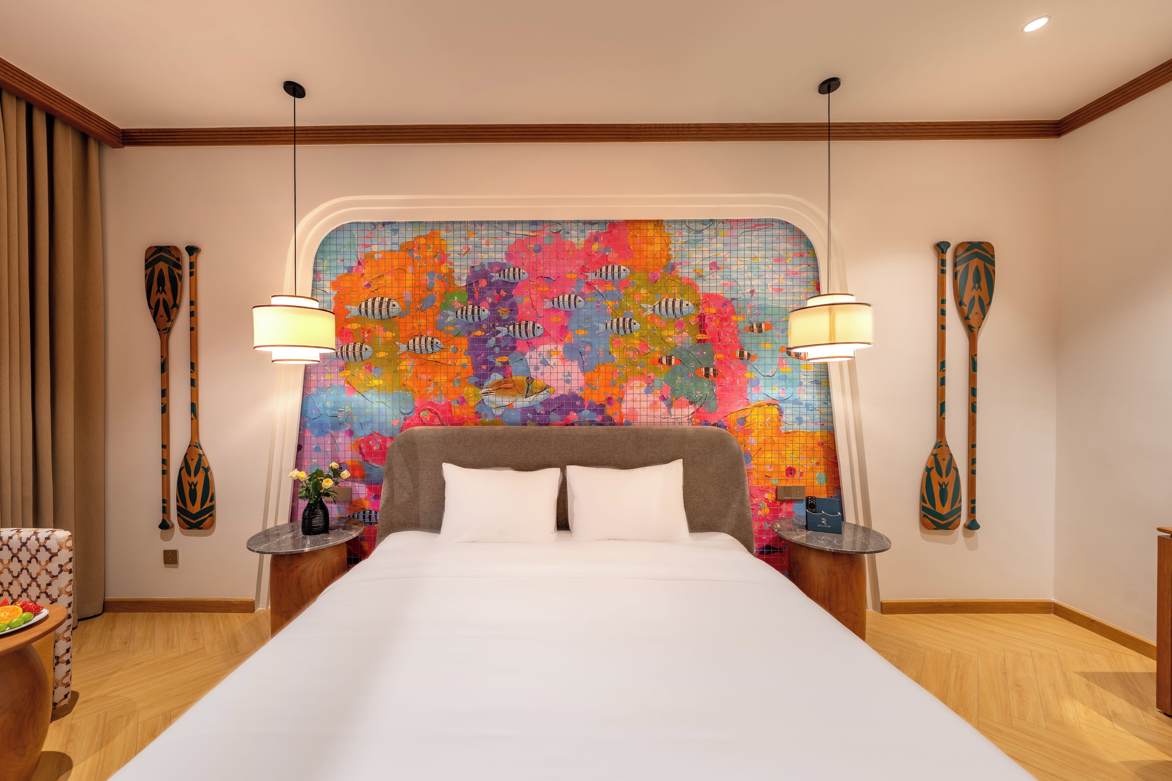 A hotel room with a large bed, a colorful wall mural, two lamps, and a wooden headboard.