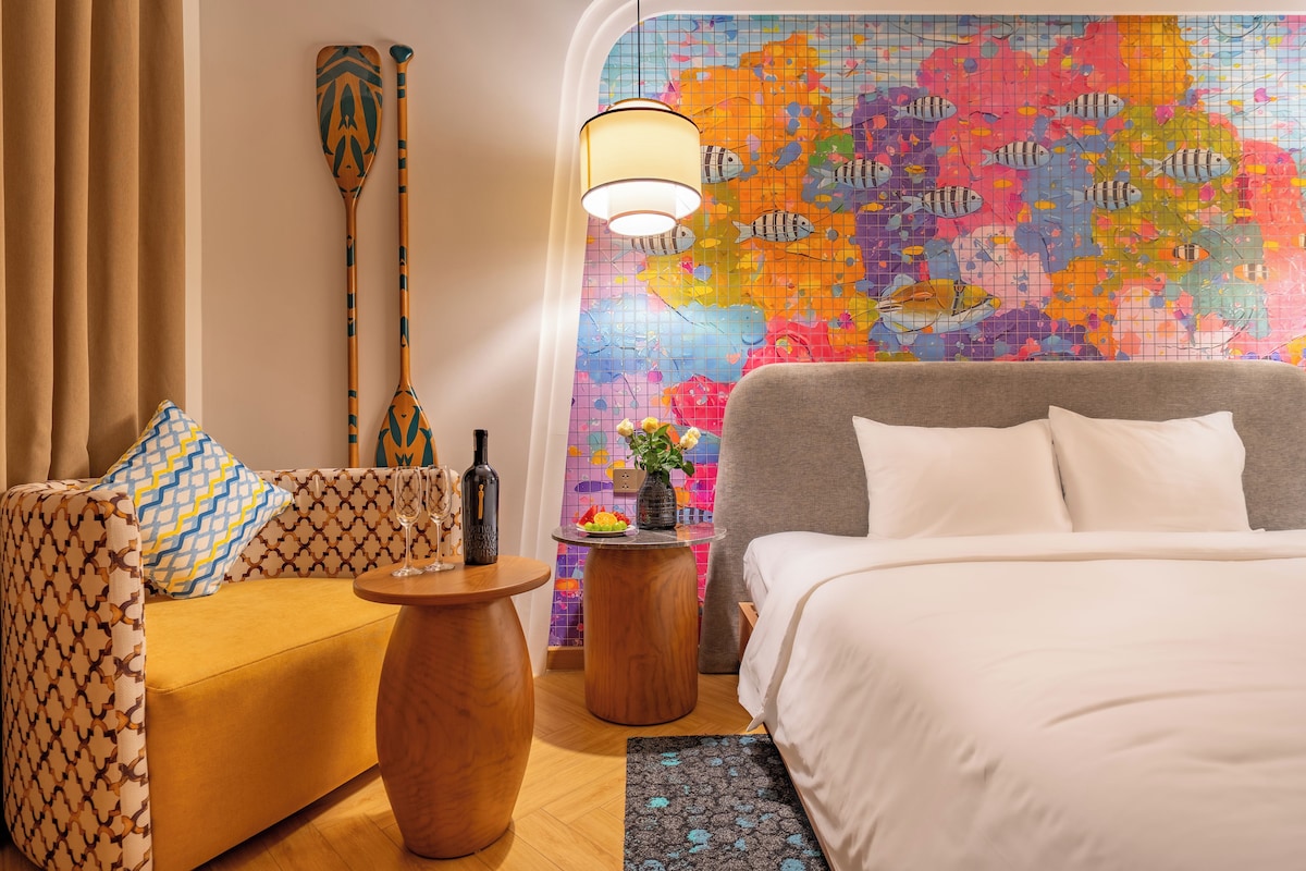A hotel room with a bed, a sofa, a small round table with a bottle and glass, a wall with a colorful fish mural, and a wooden paddle.