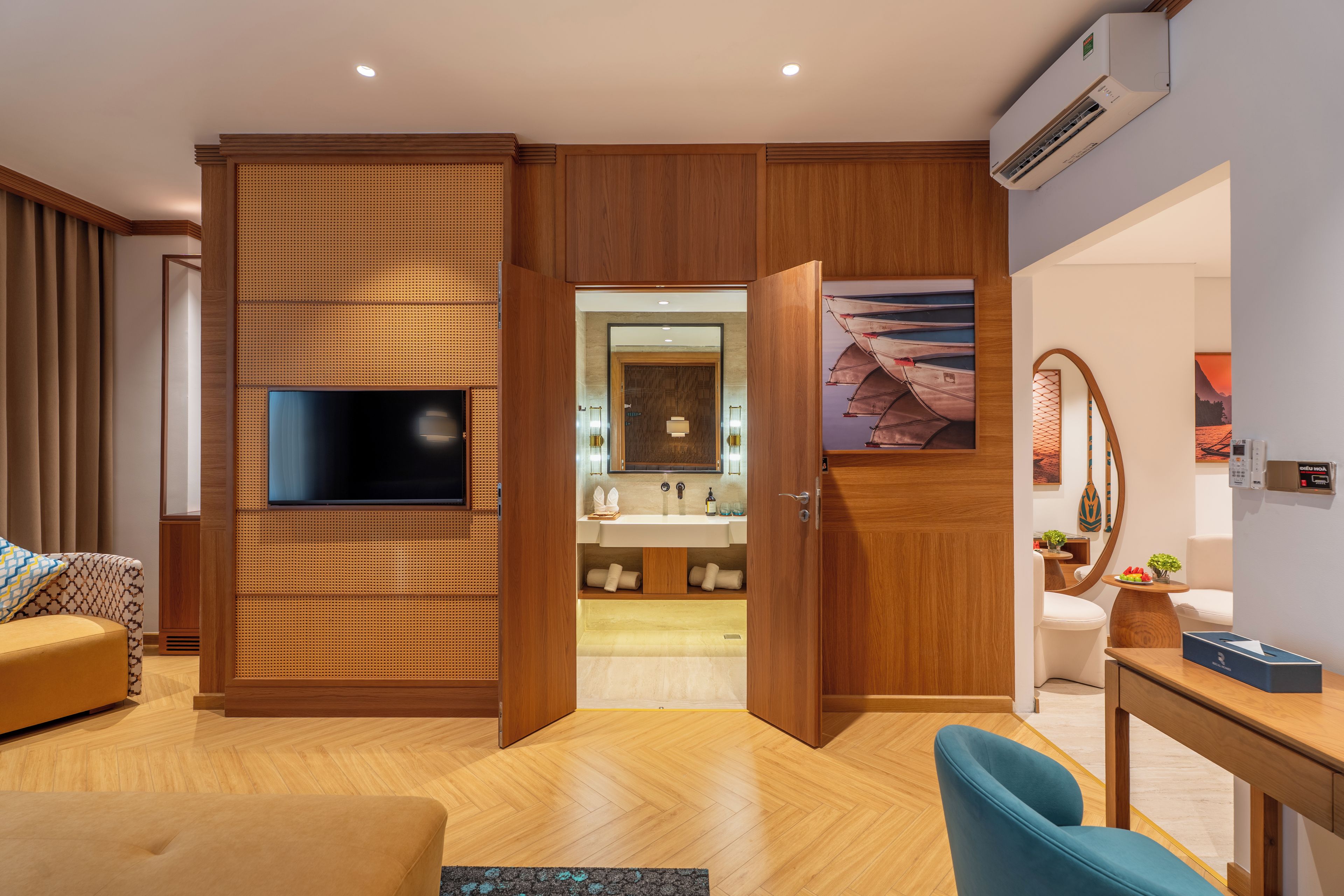Ein modernes Hotelzimmer mit Flachbildfernseher, Holzpaneelen, einem Badezimmer mit Waschbecken und Spiegel sowie einem Essbereich mit Tisch und Stühlen.