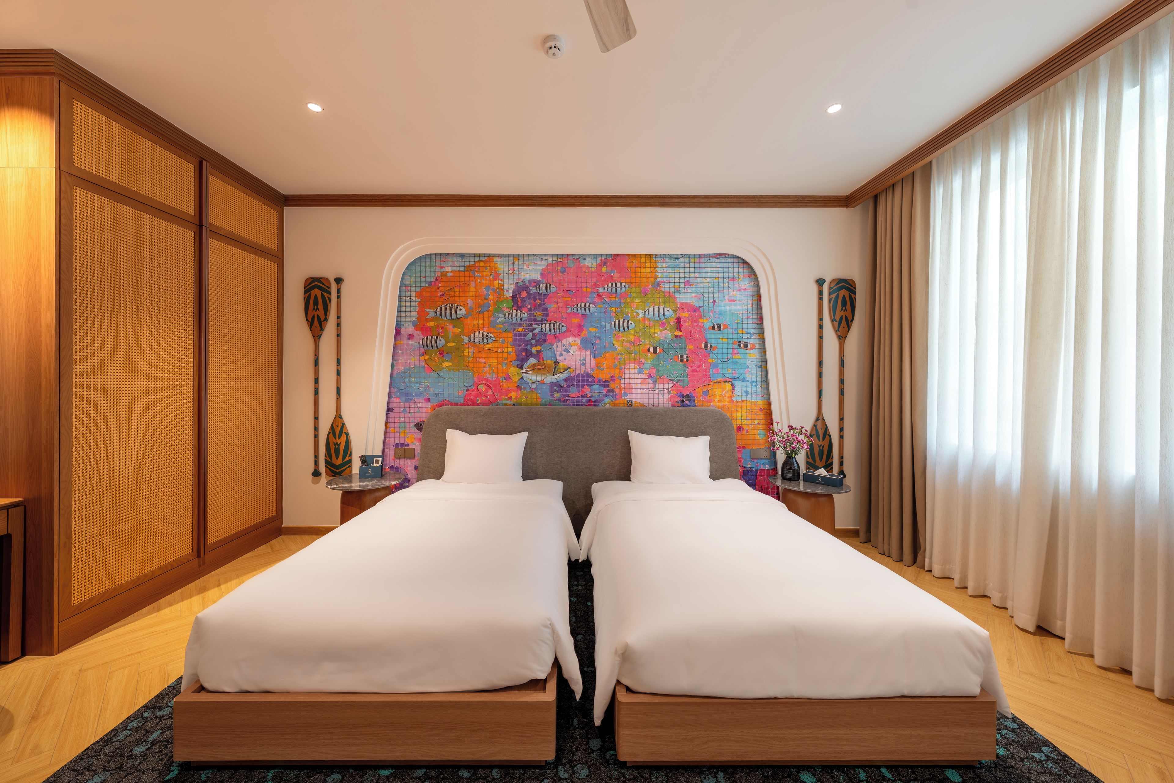 A hotel room with two beds, a large colorful map on the wall, wooden furniture, and a window with curtains.