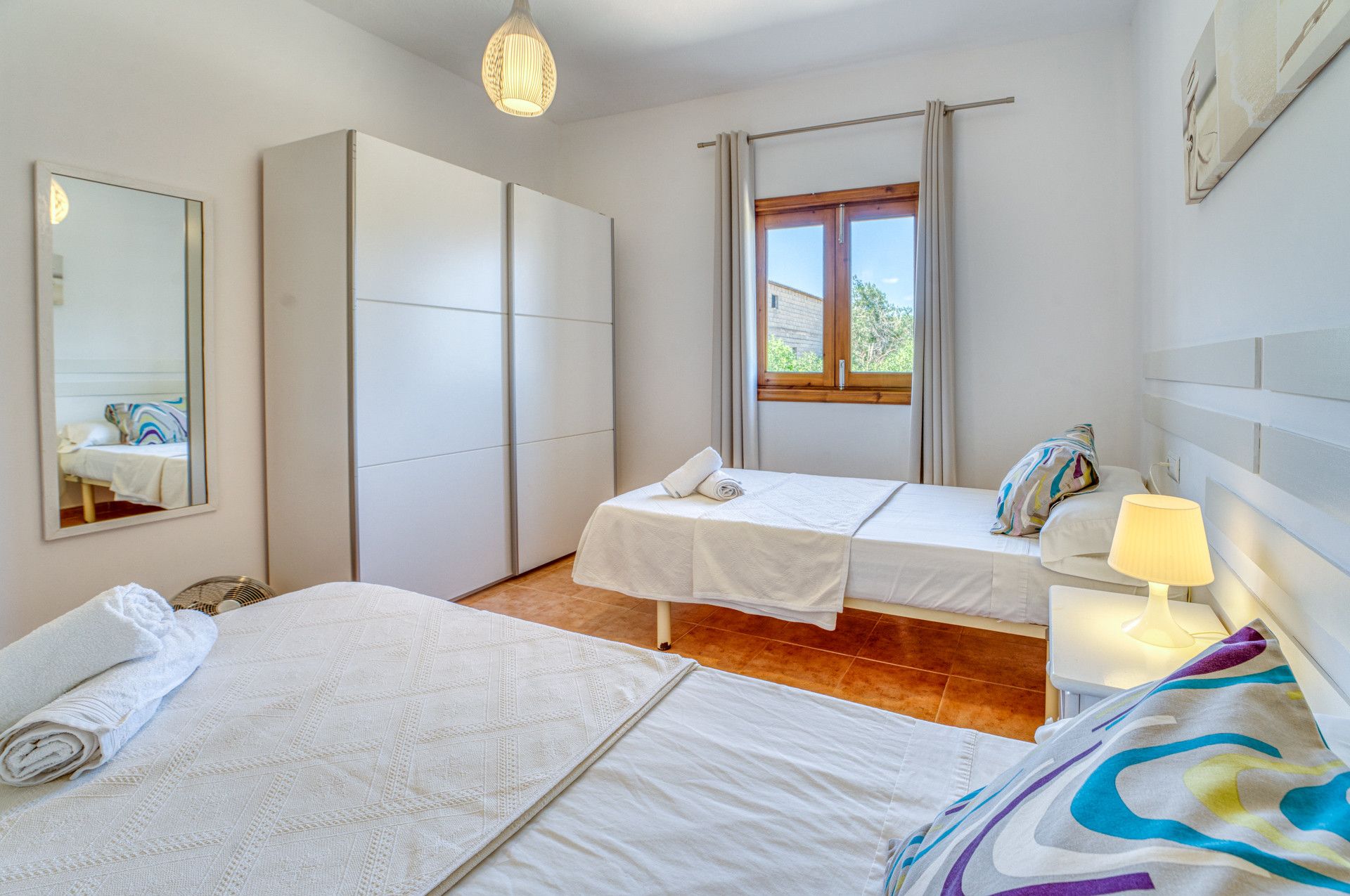 A hotel room with two beds, a wardrobe, a window with curtains, a bedside lamp, and a mirror.
