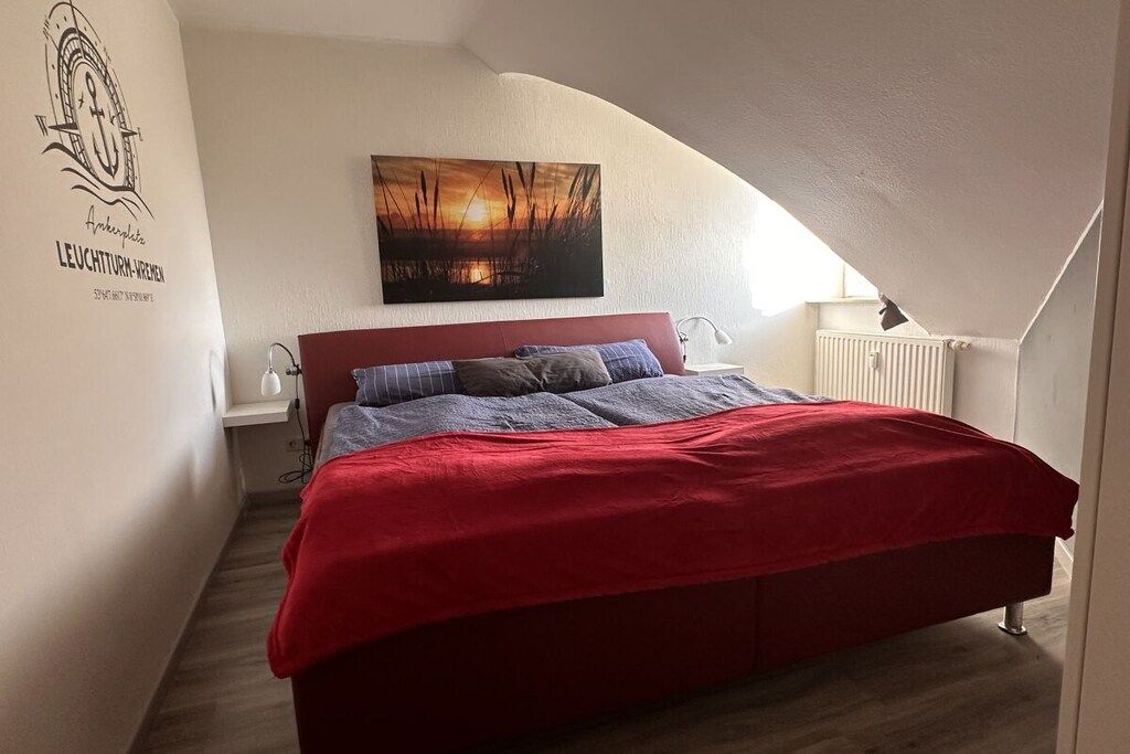 Une chambre à coucher avec une couvre-lit rouge, une table de chevet avec une lampe, une œuvre d’art accrochée au mur et un radiateur.