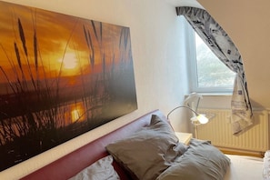 A bedroom with a bed, pillows, a nightstand, a lamp, a window with curtains, and a painting on the wall. - Holiday apartment with 1 bedroom (Wurster Nordseeküste \/ Wremen)