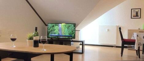 A modern living room with a television, a dining table with wine glasses, a potted plant, and a radiator.