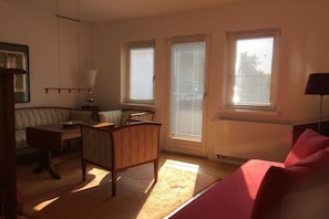 A room with a wooden desk, a red couch, and a radiator. There are three windows with blinds, and a lamp is on the desk. - Wendland-am-Weinberg (Hitzacker)