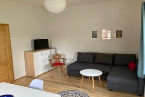 A room with a sofa, a small white table, a wooden chair, a TV on a cabinet, and framed pictures on the wall. - Holiday home in Herrnhut with barbecue (Herrnhut)