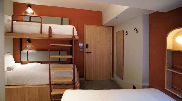 A hotel room with a bunk bed, a single bed, a door, a wall-mounted shelf, and a light fixture.