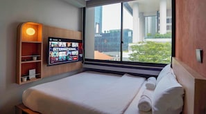 A hotel room with a large bed, a television displaying a program, a wooden shelf with a plant, and a window with a view of buildings. - ST Signature Midtown (Singapore)