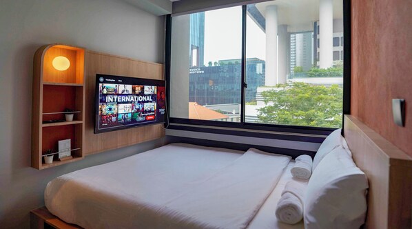 A hotel room with a large bed, a television displaying a program, a wooden shelf with a plant, and a window with a view of buildings. - ST Signature Midtown (Singapore)