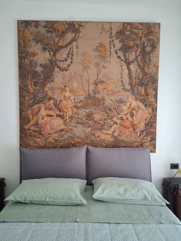 Cama com uma grande tapeçaria retratando uma cena campestre, dois travesseiros e uma mesa de cabeceira com abajur.
