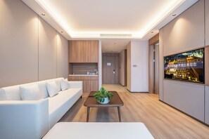 A modern hotel room with a sofa, a small table with a plant, a kitchen area, and a TV displaying a cityscape.