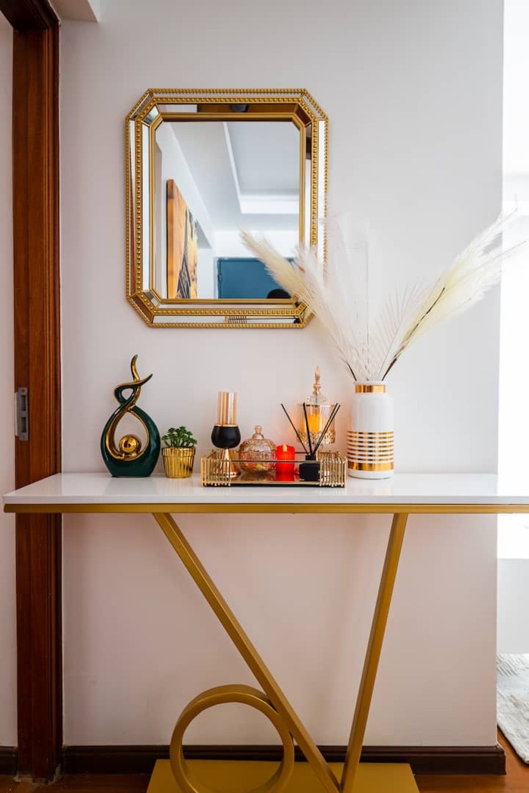 A white console table with a gold frame mirror, decorative items, and a vase with white feathers.