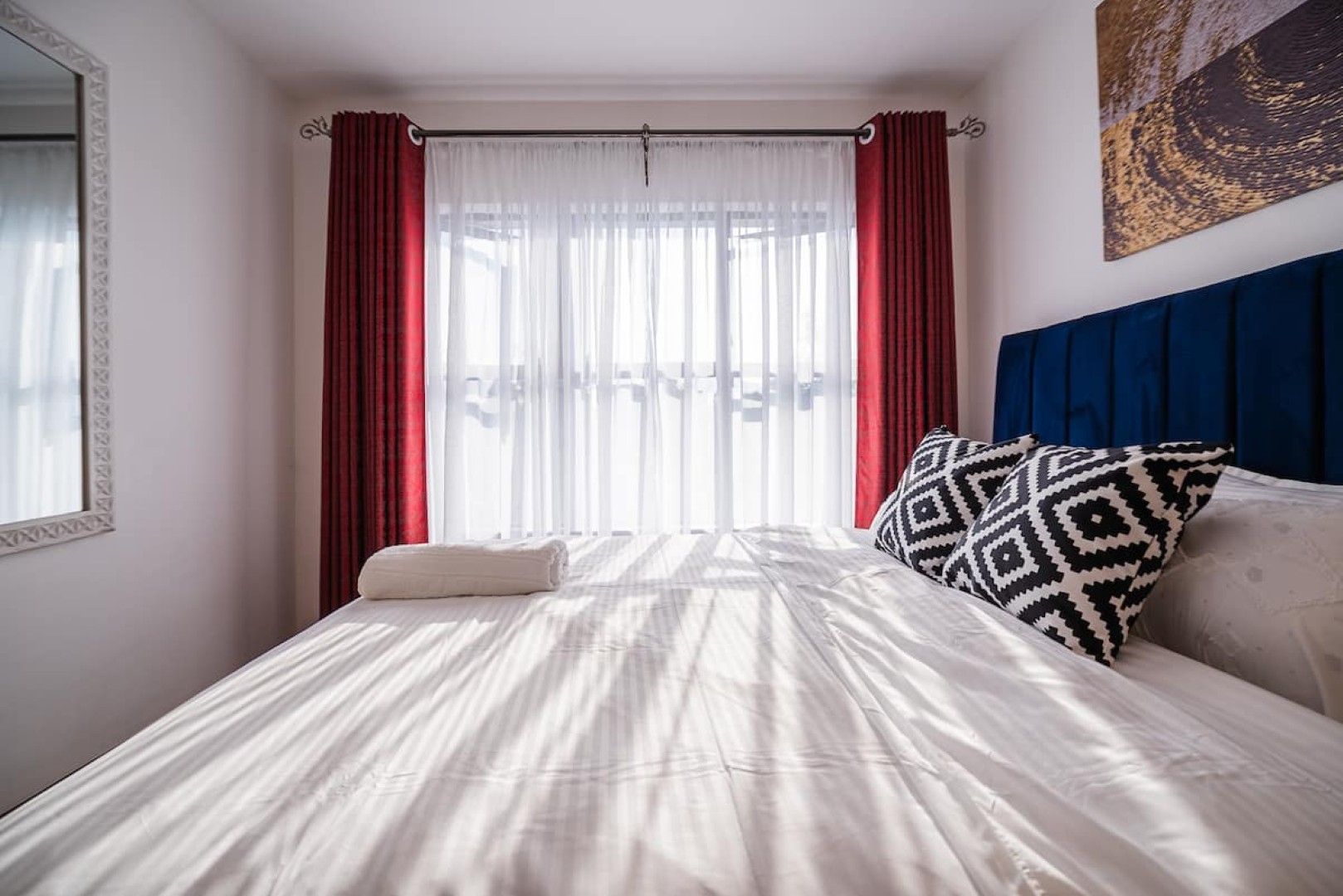 A hotel room with a bed, a window with sheer curtains, a headboard, and a wall-mounted artwork.