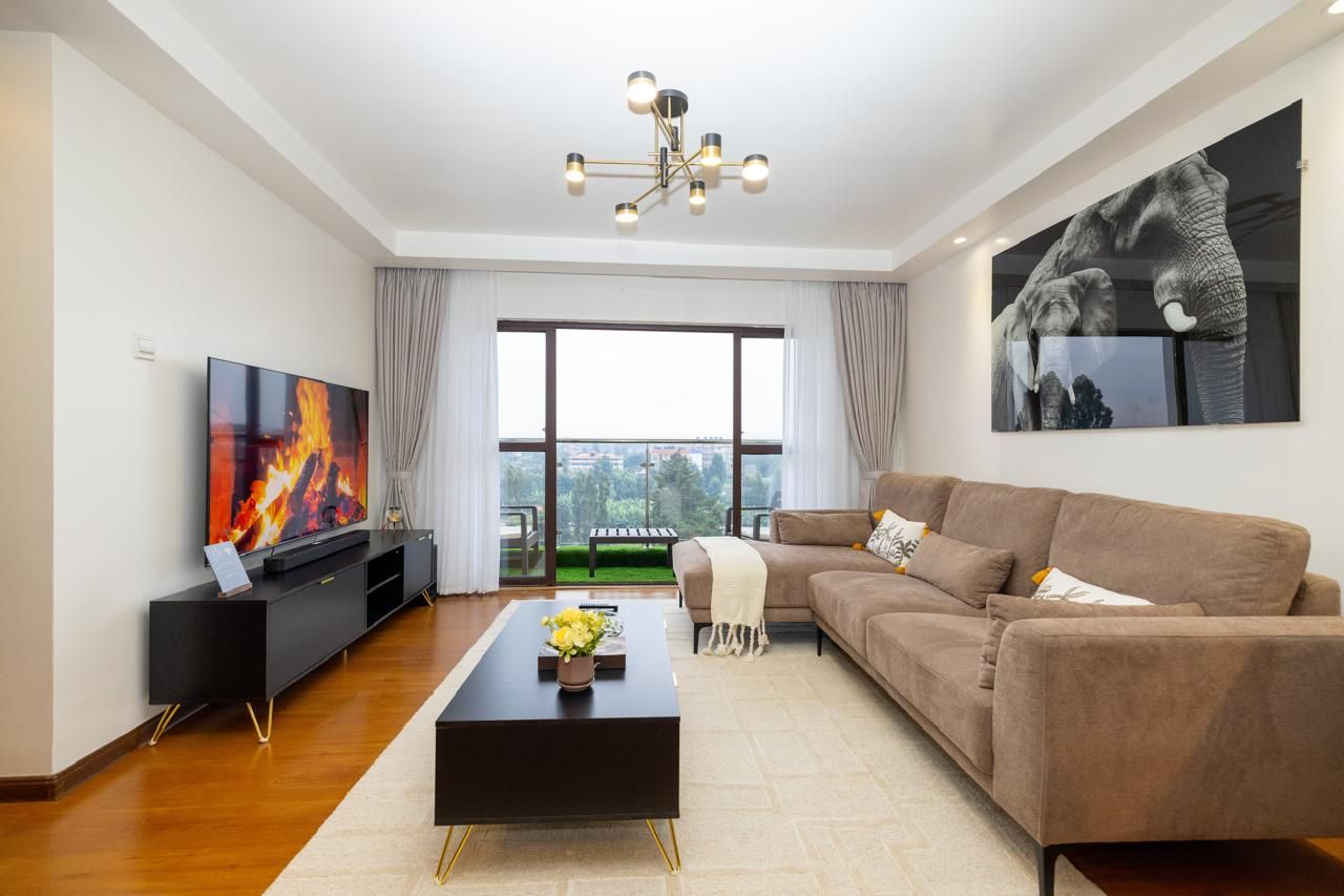 A modern living room with a flat-screen TV, a black entertainment unit, a brown sofa, a coffee table, and a large window with a view.