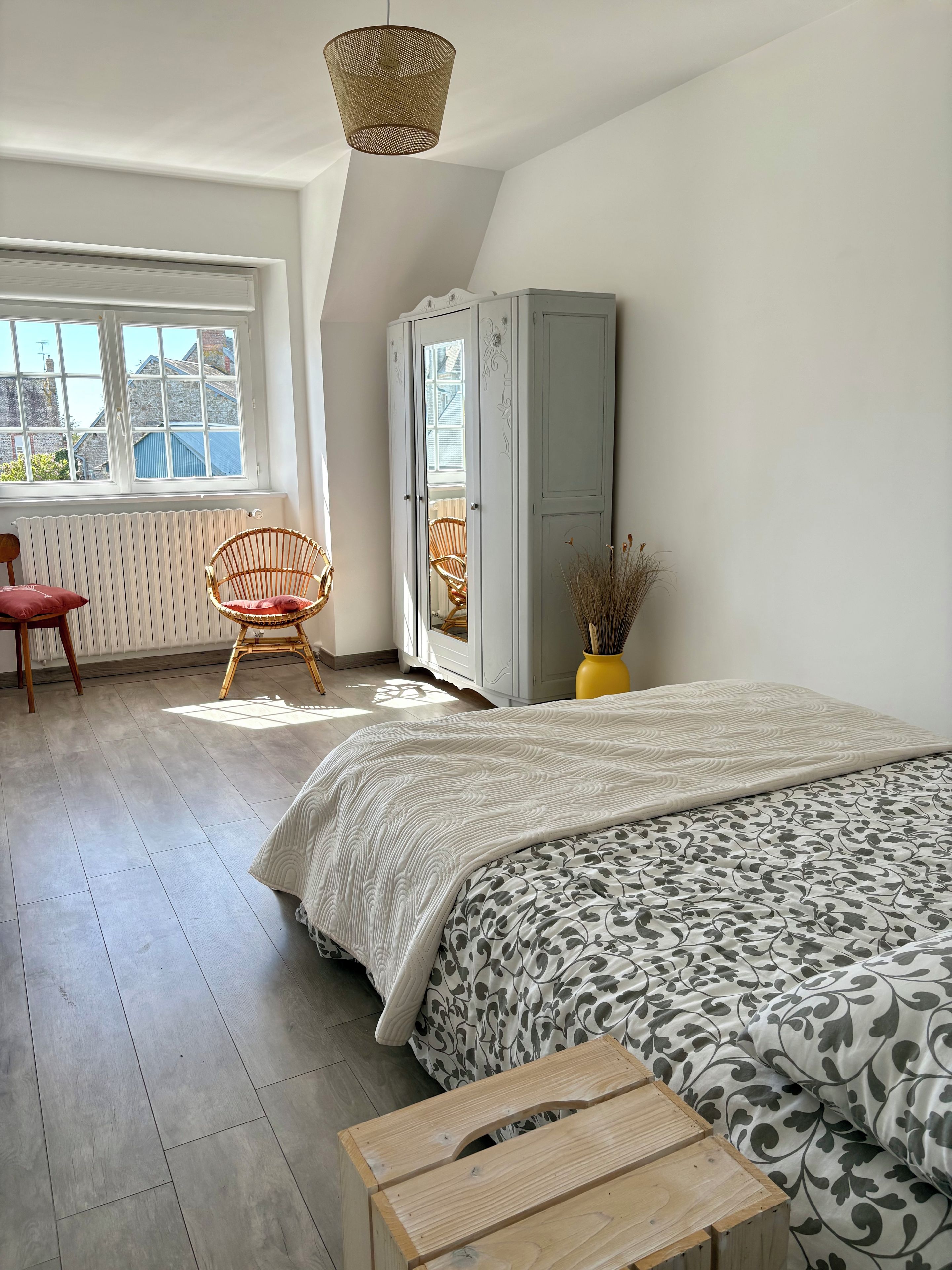 Une chambre à coucher comprenant un lit, une chaise, une armoire et une fenêtre donnant sur des immeubles.