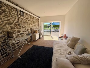 A room with a stone wall, a washing machine, a drying rack, a sofa, and a view of the outside. - My Little Farmhouse: A Quiet Family Home with an Enclosed Garden - 1.5 km from the sea 🌊 (Dragey-Ronthon)