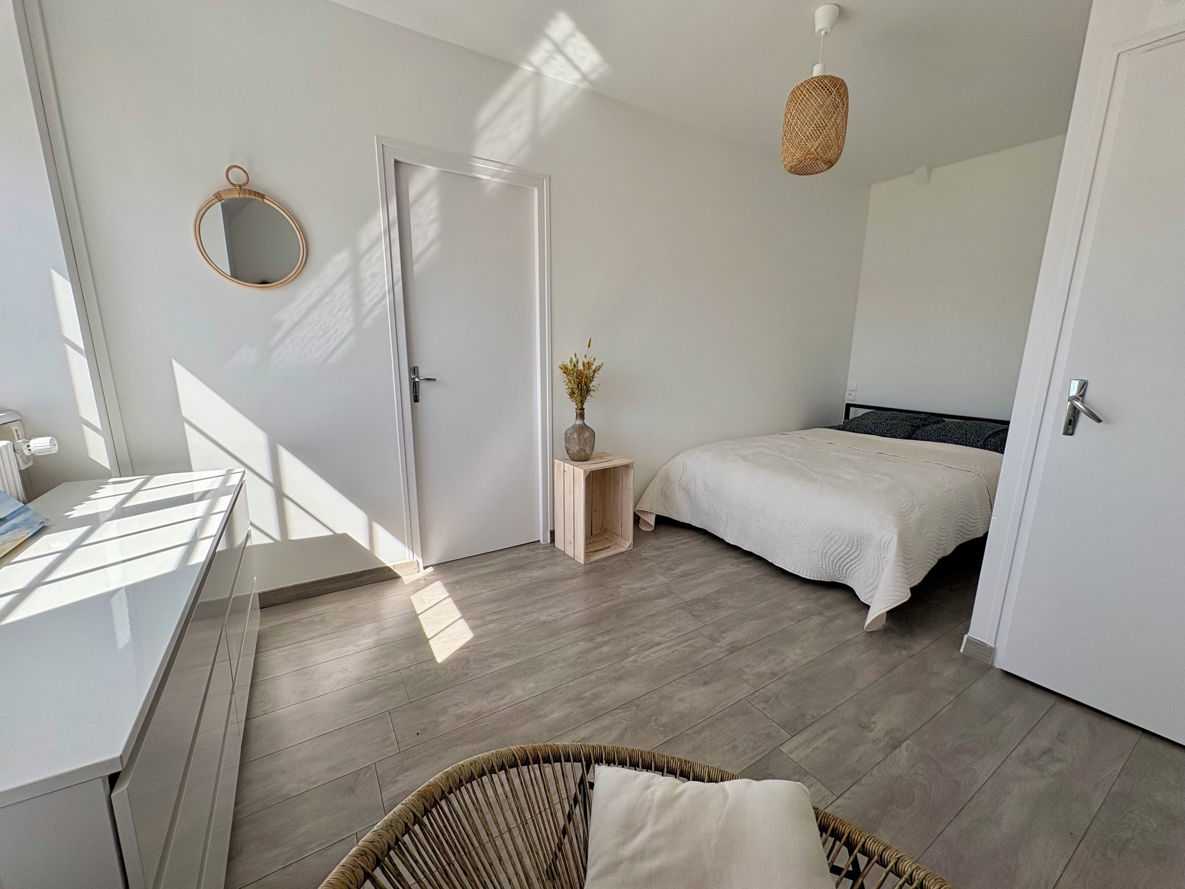 A bedroom with a bed, a bedside table with a vase, a wicker chair, and a door.