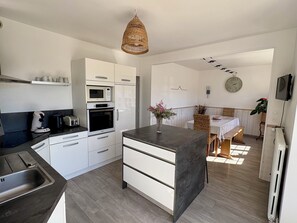 A modern kitchen with white cabinetry, a central island, and a dining area with a table and chairs. - My Little Farmhouse: A Quiet Family Home with an Enclosed Garden - 1.5 km from the sea 🌊 (Dragey-Ronthon)